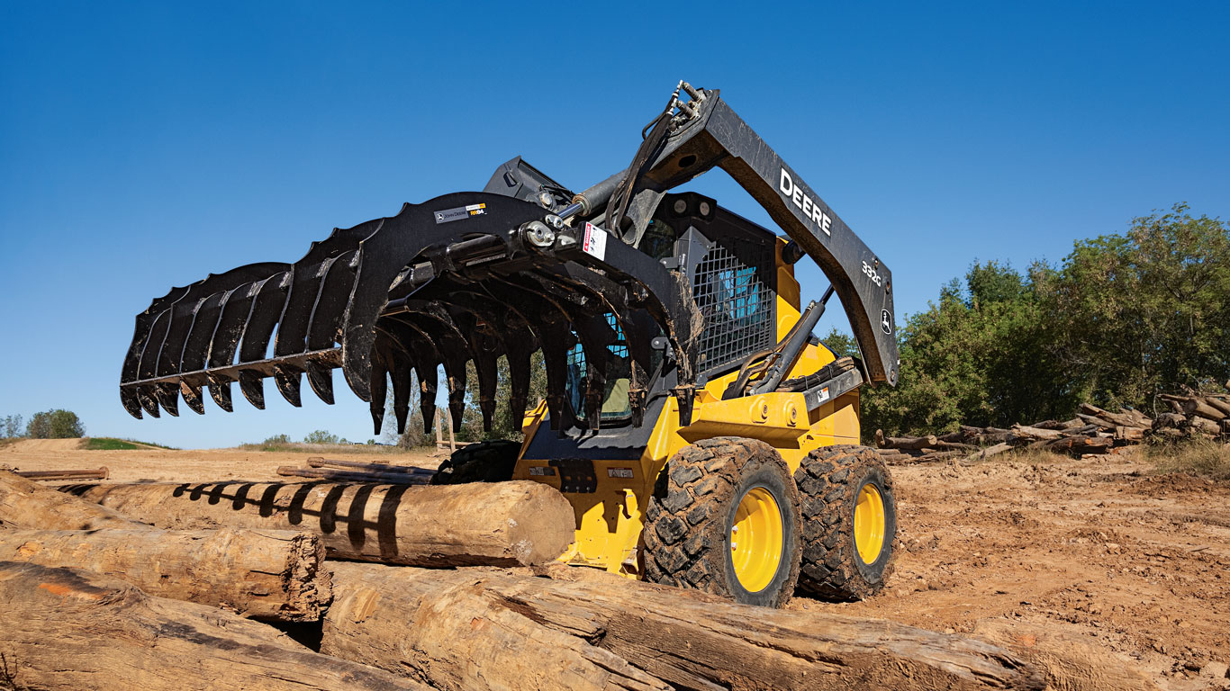 Une chargeuse à direction différentielle John Deere équipée de l’accessoire râteau dessoucheur-déracineur ramasse des grumes.